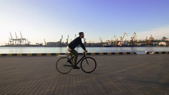 Stylish Handsome Man Riding Bicycle During Sunset or Sunrise with Sea Port on Background Slow Motion