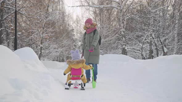 A happy mom pulls a snowcat sled with a baby in yellow warm clothes along a snowy path in the park.