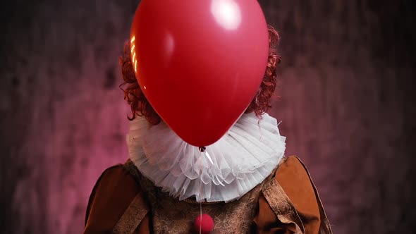 Creepy Clown Releases a Red Ball and Shows His Face. Clown in a Colorful Makeup