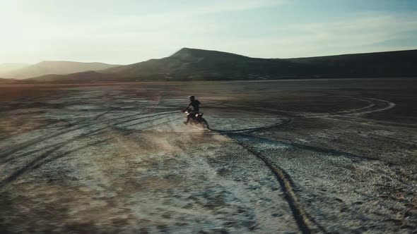 Drone View of Man on Motorbike Rides on a Dry Lake with Dust and Beautiful Hills on Background in