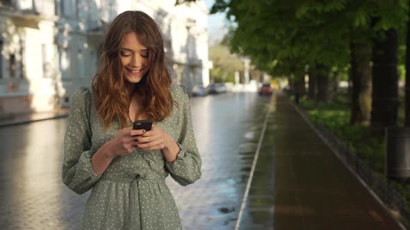 Portrait of Happy Lovely Woman Wearing Dress Smiling and Holding Mobile Phone While Walking Through