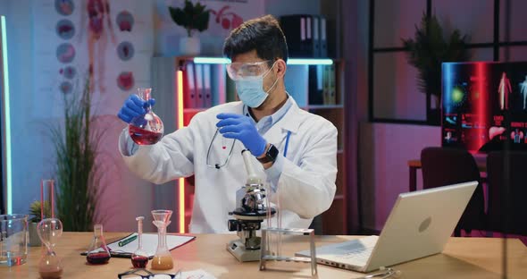 Chemist in Protective Mask and Uniform Working with Chemical Liquids in Evening Lab