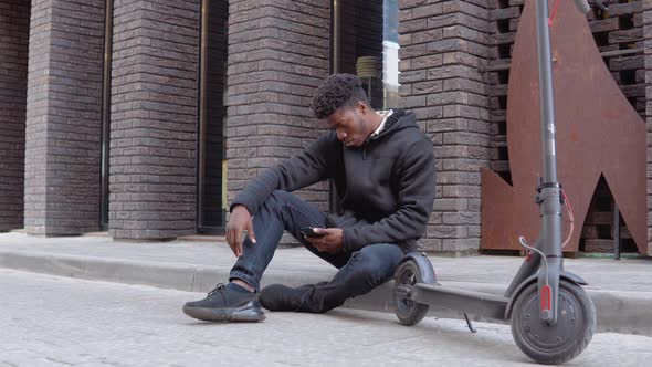 A Young African Man in a Black Sweater and Sneakers Sits on the Sidewalk Near a Building with a Dark