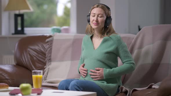 Relaxed Happy Pregnant Woman in Headphones Enjoying Music Caressing Belly and Looking at Camera