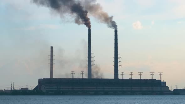 Coal Power Plant High Pipes with Black Smoke Moving Upwards Polluting Atmosphere Over Lake Water