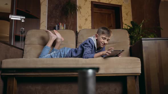 Smart Boy Lying on the Couch at Home and Watching Cartoon or Funny Video on Tablet PC