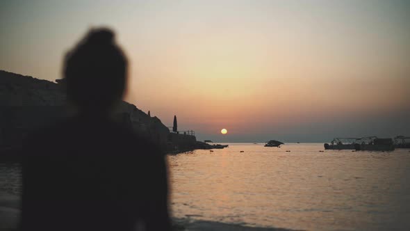 Back view of a young woman seated in front of the sunset by the sea