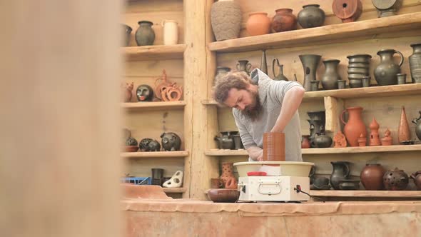 A young bearded potter works with a potter's wheel to create a tall brown jug