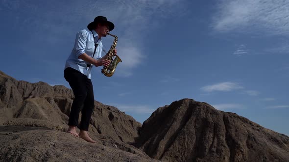 Barefoot Male Musician Plays a Wind Instrument Saxophone in the Mountains Against the Sky