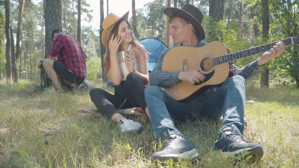 Loving Young Couple Playing Ukulele in Sunlight and Singing with Blurred Friends Camping at the