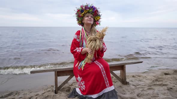 Happy Ukrainian Woman in Red Traditional Dress with Rustic Wheat Bouquet Sitting on Sandy River