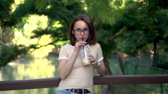 A Young Woman with Glasses Stands on a Bridge in the Park and Drinks Cold Coffee Closeup. In the