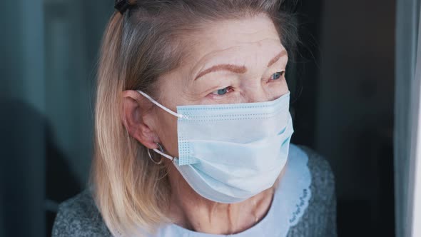 Portrait of Elderly Woman with Medical Face Mask in Quarantine Looking Through the Window