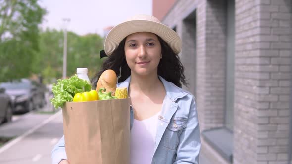 A Young Attractive Woman in a Denim Jacket and Hat Carries a Grocery Bag While Having a Good Mood