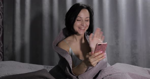 Woman Sitting on Bed Under Blanket. Enjoying Video Chat To Friend on Smartphone