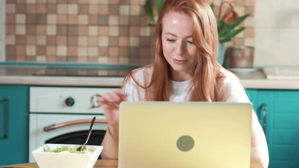 Cute woman with red hair works in the morning with a laptop in the kitchen.