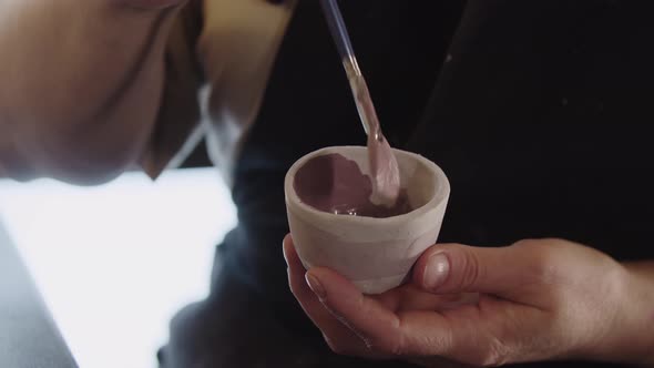 Hands of an Old Woman Painting a Little Clay Cup with Dusty Rose Color