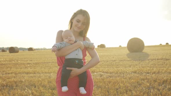 Happy Mother Wih Her Lovely Baby on Hands Smiles and Looks at Camera in Field