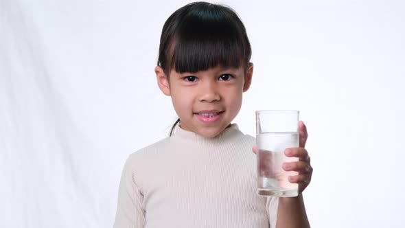 Cute little asian girl holding a glass of water and showing thumbs up on white background in studio.