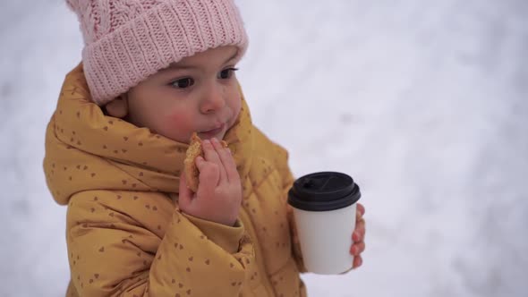 Child Girl Drinking Cocoa and Eating Cookie Outdoors in Winter Snowy Day