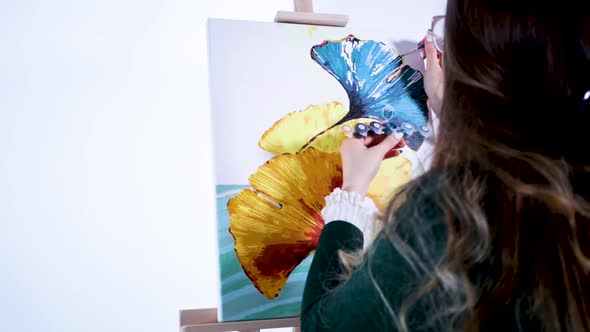 Back View Of A Young Girl Artist Painting The Complex Edges Of Ginkgo Leaf Using A Fine Paint Brush.