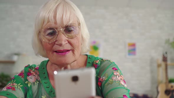 Elderly Woman with Gray Hair Uses a Smartphone Sitting at a Table in the Living Room Close Up