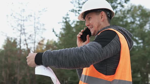 The foreman at the construction site is talking on the phone and checking the drawings.