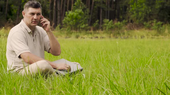 A Man with a Laptop Is Talking on the Phone on the Green Lawn