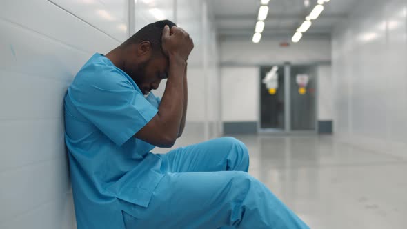 Stressed Tired Young Afro Surgeon Sitting on Floor in Hospital Corridor After Difficult Surgery