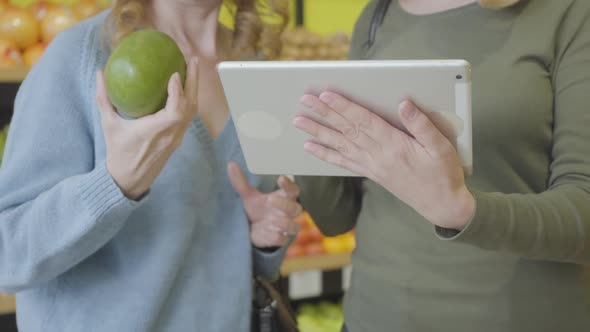 Two Unrecognizable Caucasian Women Using Tablet To Check Pomelo Origin in Grocery. Serious Ladies