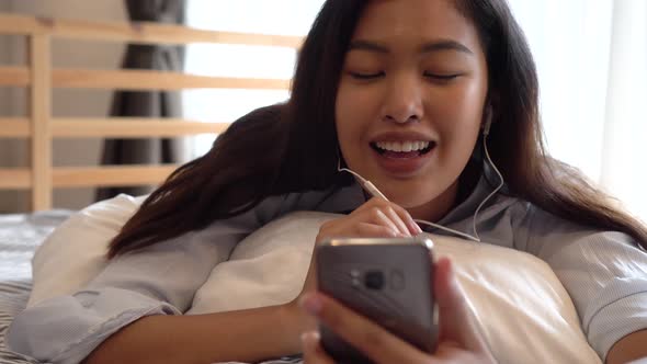 Happy Young Asian Girl Lying Down on Bed While Making a Video Call with Smartphone & Waving at