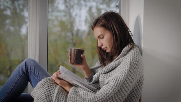 Portrait of a Brunette Girl Reading a Book at the Window with a Cup of Hot Tea