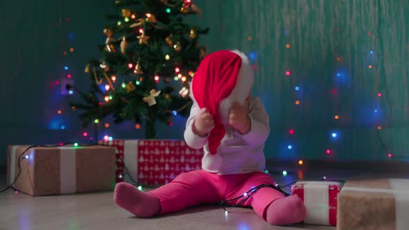 A funny little girl sits near the Christmas tree and takes off her Christmas hat