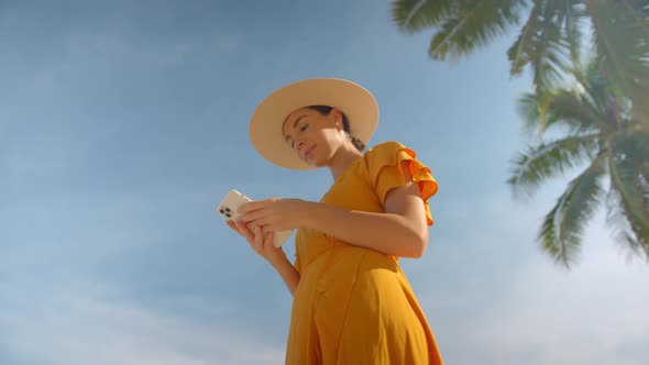 Lovely Woman in Summer Yellow Dress and Straw Hat Standing Under Palmtree and Using Her Cell Phone