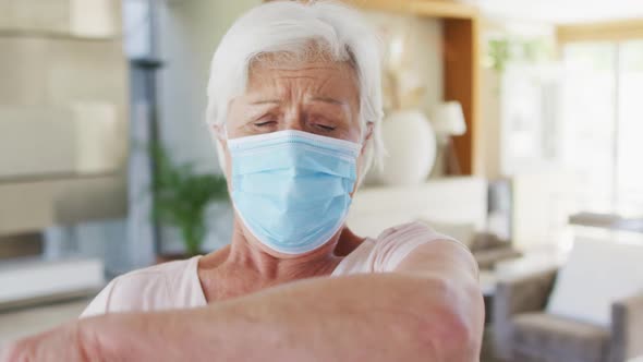 Senior caucasian woman wearing face mask sneezing on her elbow at home