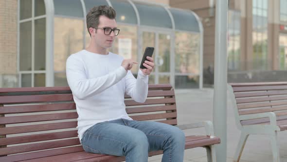 Young Man Celebrating Online Success on Smartphone While Sitting Outdoor on Bench