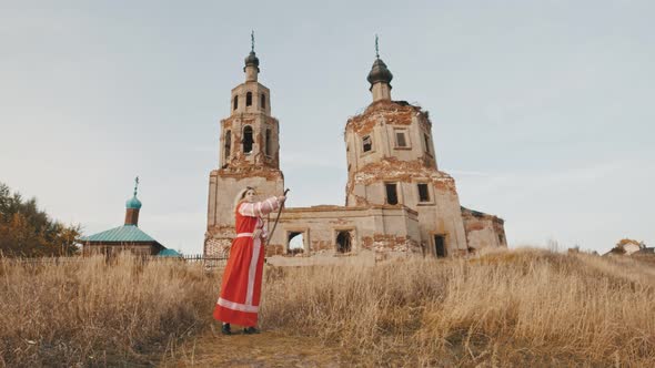 Woman in Red Dress Wields Swords Against the Background of a Ruined Old Church