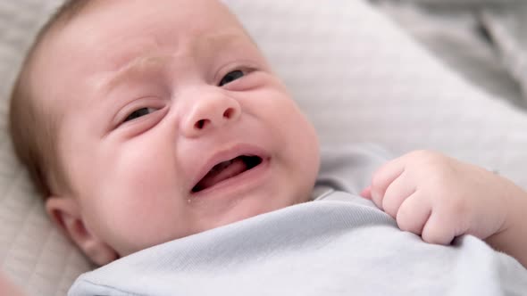 Dissatisfied Crying Upset Sad Newborn Baby Boy Lying in Nursery Cocoon with Blue Clothes on Bed in