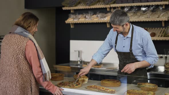 Happy Bakery Salesman Cuts a Piece of Cake for a Female Customer and They Have a Casual Conversation