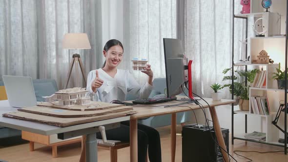 Smiling Asian Woman Engineer Holding House Model And Showing Thumbs Up Gesture To The Camera