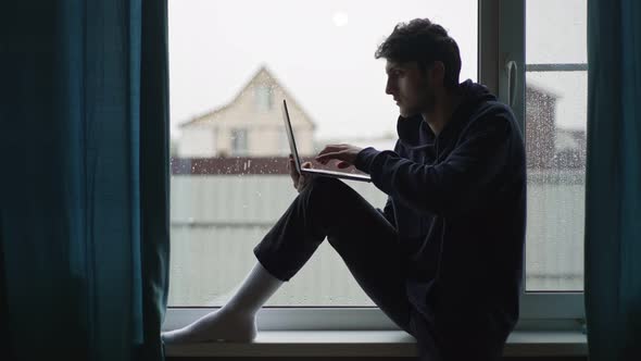 A Young Man is Typing on a Laptop Sitting at the Window Against the Backdrop of a Picturesque Rural