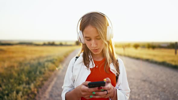 Young Girl Stands on Country Road in Field and Uses Her Phone to Switch Songs in Her Headphones