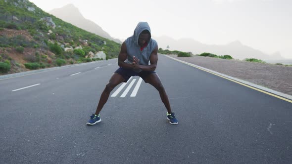 African american man wearing a hoodie performing stretching exercise on the road