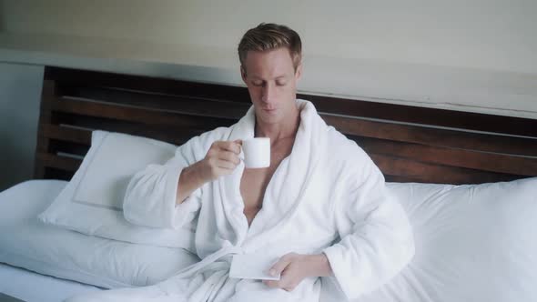 Young Man in Bathrobe Drinks Coffee in Bed Near Laptop