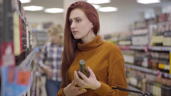 Side View of Young Beautiful Redhead Woman Examining Shelves with Alcohol and Bottle of Red Wine in