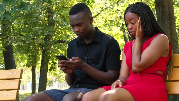 A Black Woman Is Frustrated That a Black Man Is on His Smartphone As They Sit on a Bench in a Park