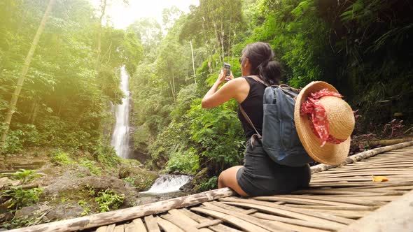 Young Traveler Girl Sitting on Wooden Bridge and Taking Photos of Jungle Waterfall