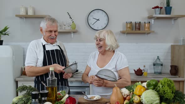 Senior Woman and Man Making a Funny Dance with Strainers. Dancing While Cooking Together in Kitchen