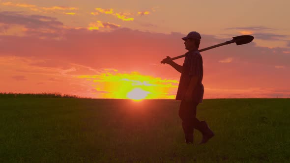 Silhouette of the Man with the Shovel Walking Through the Field at Sunset