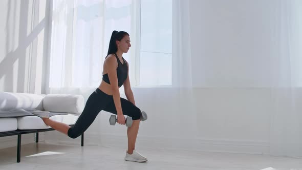 Brunette in Black Sportswear in a White Apartment Makes a Split Squat with Dumbbells in Her Hands